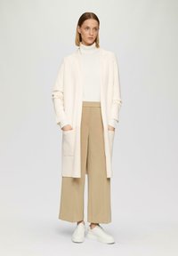 Langer, hellrosa Cardigan mit offenem Vorderteil und Taschen, getragen über einem weißen Rollkragenpullover und beigen weit geschnittenen Hosen, kombiniert mit weißen Sneakers.