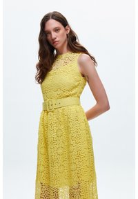 Das gelbe Spitzenkleid mit Blumenmustern hat einen ärmellosen Schnitt, eine taillierte Taille mit einem passenden Gürtel und einen schwingenden Rock bis zur Mitte der Wade.