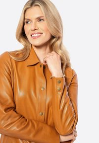 Bruine leren jas met drukknoopjes en kraag; gladde textuur. Model poseert, houdt de kraag met één hand vast en draagt een armband.