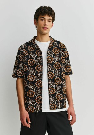 Junger Mann trägt ein schwarzes kurzärmeliges Hemd mit großen orange-weißen Blumenmuster über einem weißen T-Shirt und schwarzen Hosen.