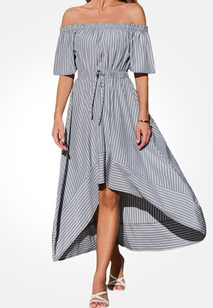 Vrouw draagt een grijs-wit gestreepte off-shoulder jurk met een elastische taille en asymmetrische zoom, gecombineerd met witte sandalen met bandjes.