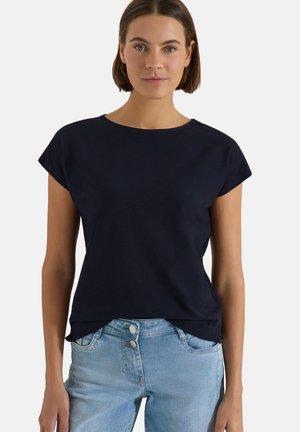 Frau mit kurzen braunen Haaren, die ein schwarzes kurzärmeliges Oberteil und hellblaue hoch taillierte Jeans trägt, vor einem schlichten hellen Hintergrund.