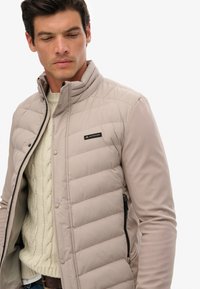 Doudoune beige clair avec col en tricot, poche zippée et détail de logo noir. Présente une texture matelassée et un tissu de manches lisse.