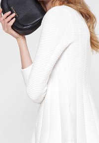 Robe blanche texturée avec un design côtelé, associée à un sac en cuir noir incurvé avec un accent doré.