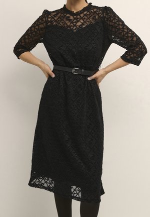 Kobieta ubrana w czarną midi koronkową sukienkę z rękawami do połowy przedramienia i czarnym paskiem, z rękami opartymi na biodrach na jednolitym tle.