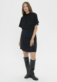 Vestido corto negro con cintura ajustada, que presenta un escote alto y mangas de volantes. Decorado con pequeños lunares blancos.