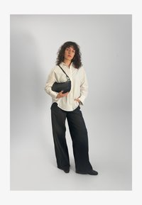 Camisa blanca abotonada, pantalones negros de pierna ancha, bolso de cuero negro con correa y zapatos negros. Pelo rizado, fondo neutro.