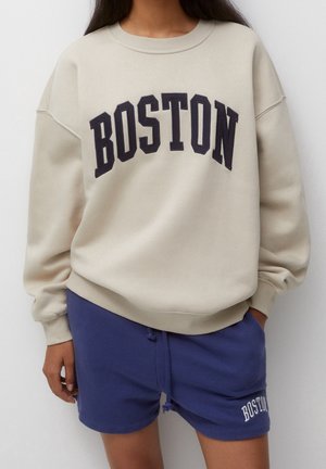 Person trägt einen beigen Pullover mit "BOSTON" in großen Buchstaben und blaue Shorts mit einem kleinen "BOSTON" Logo am linken Bein.