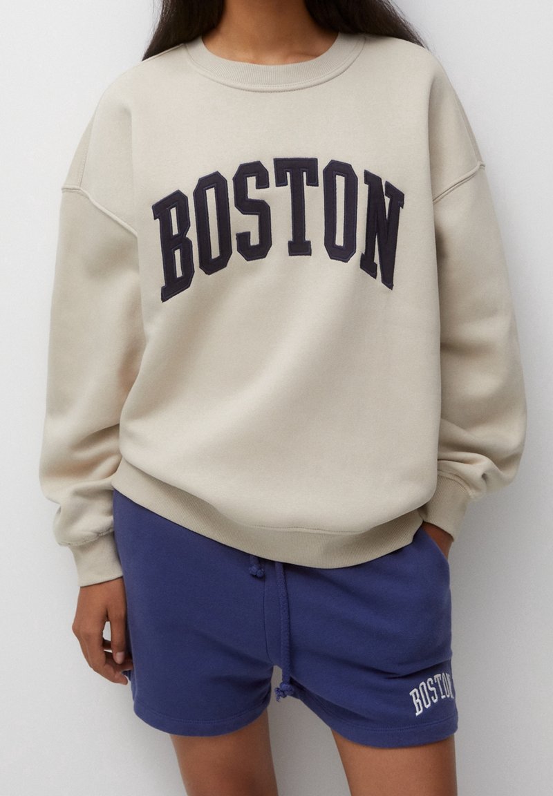Personne portant un sweat-shirt beige avec "BOSTON" en grandes lettres et un short bleu avec un petit logo "BOSTON" sur la jambe gauche.