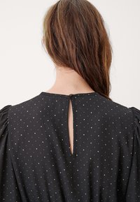 Žena s dlouhými hnědými vlasy, oblečená v černých šatech s malými bílými puntíky a knoflíkovým rozparkem na zadním výstřihu.