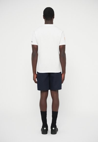 Λευκό κοντομάνικο μπλουζάκι με στρογγυλή λαιμόκοψη, συνδυασμένο με ναυτικά σορτς. Μαύρες κάλτσες και μαύρα παπούτσια με λευκά φύλλα συμπληρώνουν την εμφάνιση.