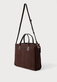 Sötétbarna velúr táska fekete bőr fogantyúkkal és állítható vállpánttal, fehér háttér előtt bemutatva.