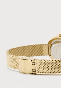 Złota siatkowa metalowa bransoleta zegarka z zapięciem wygrawerowanym "Calvin Klein" na jednolitym białym tle.