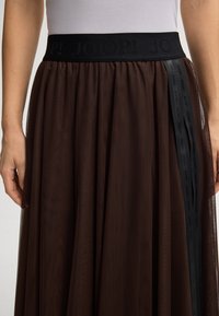 Saia de tule castanha com cintura elástica preta e detalhe de fita com logotipo vertical, usada com uma blusa branca por dentro, mãos relaxadas ao lado.