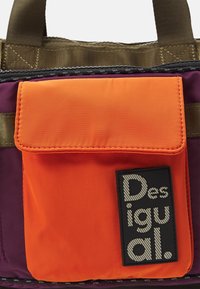 Gros plan d'un sac violet et orange avec une poche frontale orange et une étiquette noire indiquant "Desigual." Des poignées marron sont visibles en haut.