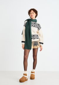 Brede gebreide trui in crème met zwarte geometrische patronen, gecombineerd met een groene sjaal, een spijkerrok en tan laarzen met witte schapenvacht accenten.