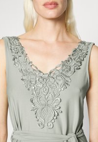 Vestido sem mangas em cinza claro, com um intrincado bordado prateado ao redor do decote em V, feito de um tecido texturizado e um laço na cintura.