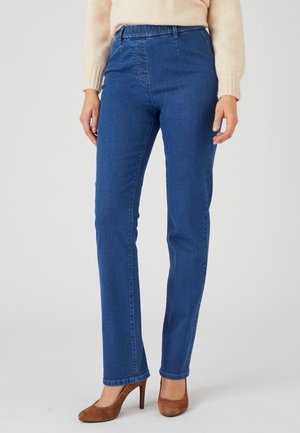 Frau trägt blaue High-Waist-Jeans und beigen Pullover, steht mit braunen High Heels sichtbar vor hellem Hintergrund.