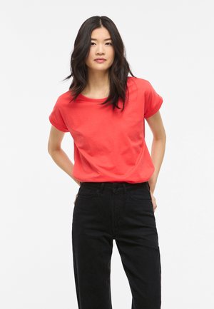 Femme aux longs cheveux foncés porte un T-shirt rouge vif à manches courtes et un pantalon noir taille haute, debout les mains derrière le dos.