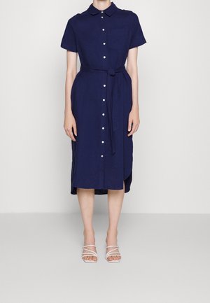 Robe chemise bleu marine à boutons, avec col, manches courtes, une poche avant et une taille nouée. Portée avec des sandales blanches à brides.