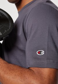 T-shirt gris à manches courtes avec un petit logo brodé sur la manche ; le tissu semble lisse et respirant.