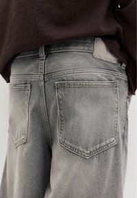 Vue arrière d'une personne portant un jean en denim gris délavé et un haut ample marron foncé, mettant en valeur les grandes poches arrière et les détails de la taille.