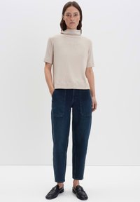 Donna in piedi che indossa un top corto a collo alto beige, jeans a vita alta blu scuro con tasche davanti e mocassini neri su sfondo bianco.