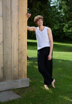 Hvid tanktop og løse sorte bukser, står barfodet ved en trævæg i en græsplæne med træer i baggrunden.
