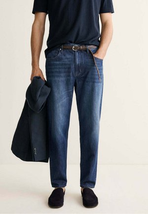 Hombre que lleva jeans azul oscuro, cinturón negro, camisa negra y mocasines negros, sosteniendo un blazer oscuro en su mano izquierda.