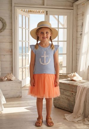 Junges Mädchen mit geflochtenen Haaren, das einen Strohhut, ein ärmelloses gestreiftes Oberteil mit Anker, einen orangefarbenen Tüllrock und Sandalen trägt, in einer von Sonnenlicht durchfluteten Strandhütte.