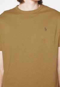 Hnědé bavlněné tričko s kulatým výstřihem, krátkými rukávy a malým zeleným vyšitým logem na levé části hrudi.