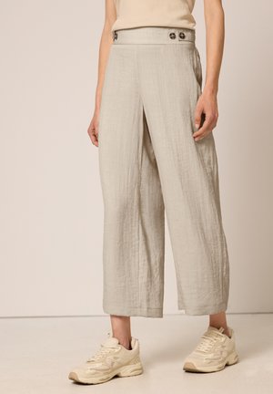 Frau trägt hellbeige, weit geschnittene, kurze Hosen mit Knöpfen vorne am Bund und cremefarbene Sneaker, steht vor einfarbigem Hintergrund.