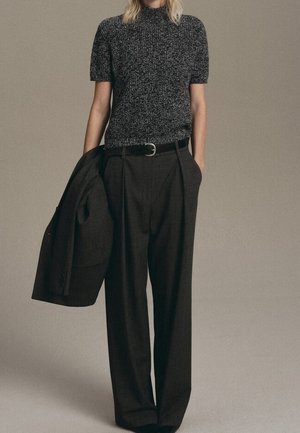 Personne portant un haut en maille à manches courtes gris foncé, rentré dans un pantalon noir ample à taille haute, tenant une veste noire assortie dans une main.