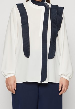 Blouse blanche à manches longues avec détails de ruban plissé noir vertical et boutons, associée à un pantalon bleu marine foncé, portée par une personne.