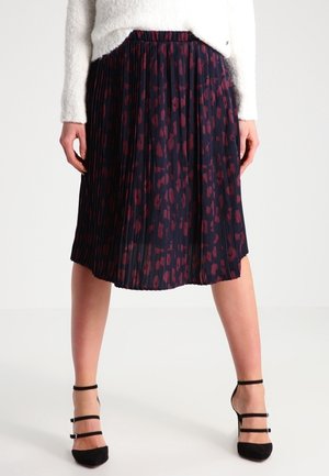Jupe plissée longueur genou avec motif abstrait bleu foncé et rouge, portée avec un pull blanc et des talons hauts noirs à brides.