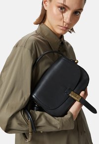 Borsa in pelle nera con un flap curvo, dettagli in ottone e una tracolla removibile, tenuta vicina a una camicia verde oliva.