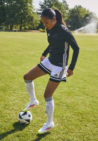 Schwarz langärmliges Sportshirt mit weißen Streifen, kombiniert mit weißen Shorts mit schwarzen Akzenten und weißen Fußballschuhen mit pinken Details.