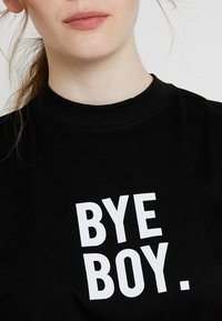 Črna bombažna majica s poudarjenim belim besedilom, ki pravi "BYE BOY." Majica s krojnim ovratnikom, kratkimi rokavi in gladko teksturo.