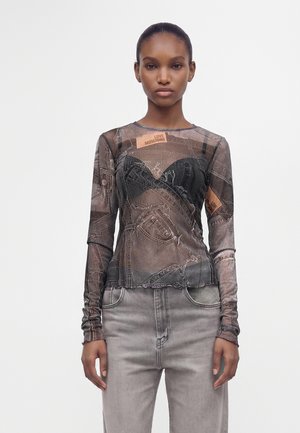 Femme portant un haut en mesh à manches longues avec du denim imprimé et des patchs "Love Moschino" sur un jean gris clair sur un fond uni.