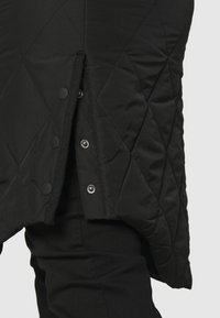 Svart kviltad jacka i sidovy med tryckknappar delvis öppna över svarta byxor.