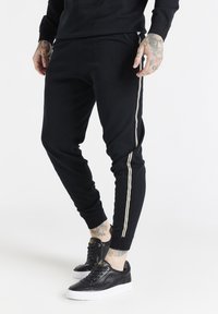 Svarta joggerbyxor med elastisk midja, med gyllene sidosträck och en diskret präglad logotyp. Matchas med svarta sneakers.