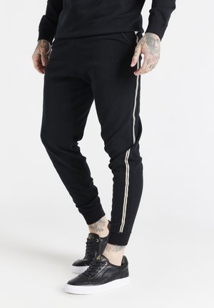 Schwarze Jogginghosen mit einem elastischen Bund, die goldene Seitestreifen und ein dezentes geprägtes Logo aufweisen. Kombiniert mit schwarzen Turnschuhen.