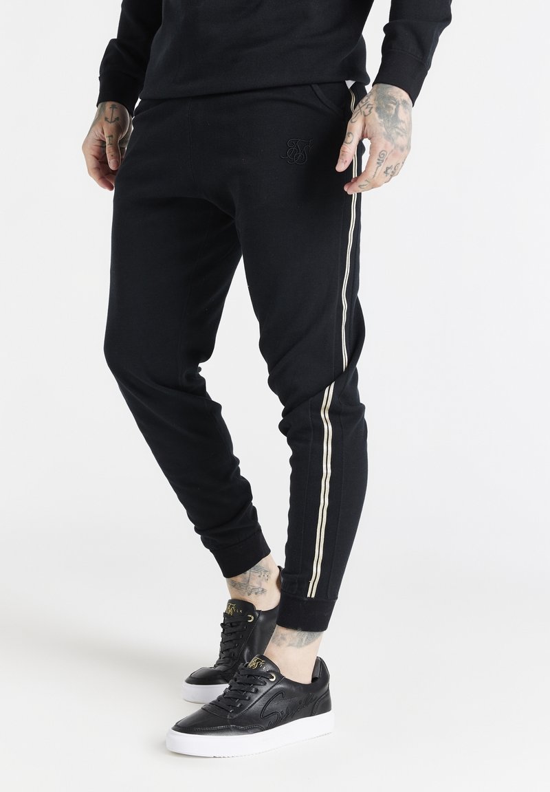 Svarta joggerbyxor med elastisk midja, med gyllene sidosträck och en diskret präglad logotyp. Matchas med svarta sneakers.