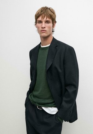 Giovane uomo con capelli castani corti che indossa un blazer scuro, maglione verde, camicia bianca e pantaloni neri su uno sfondo chiaro e semplice.