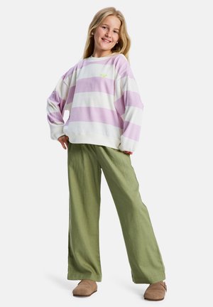 Lekka bluza z długim rękawem w białe i liliowe paski, z małym zielonym logo. Połączona z luźnymi, zielonymi spodniami i beżowymi butami.