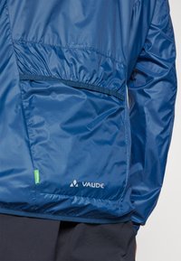 Navy blue padded jacket with a smooth texture, elastic waistband, zippered side pocket, and a visible VAUDE logo at the hem.