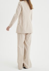 Traje beige a medida con blazer estructurado que presenta botones en las mangas y pantalones de corte relajado. Tejido suave con mínimas arrugas.