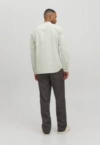 Camisa de manga larga verde claro con un corte relajado, puños abotonados y dobladillo redondeado, combinada con pantalones rectos gris oscuro y zapatos claros.