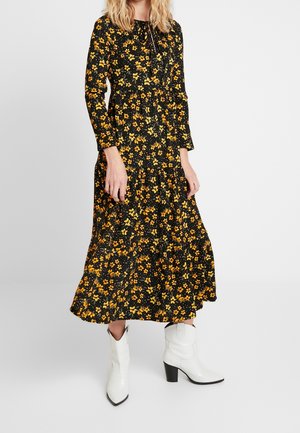 Woman wearing a long black dress with orange and yellow floral print and white mid-calf heeled boots, standing against a plain background.
