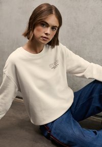 Felpa crema con il testo "CITY CLUB" in ricamo sottile, abbinata a jeans blu larghi con una striscia rossa laterale. Sfondo grigio con texture.